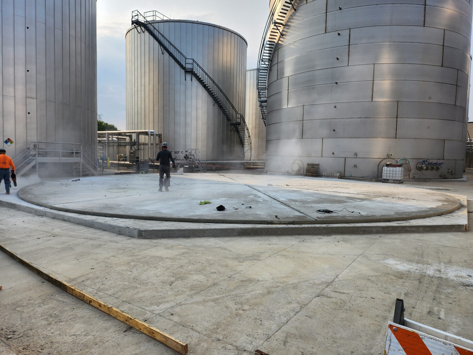 Workers cleaning concrete floor near industrial silos.
